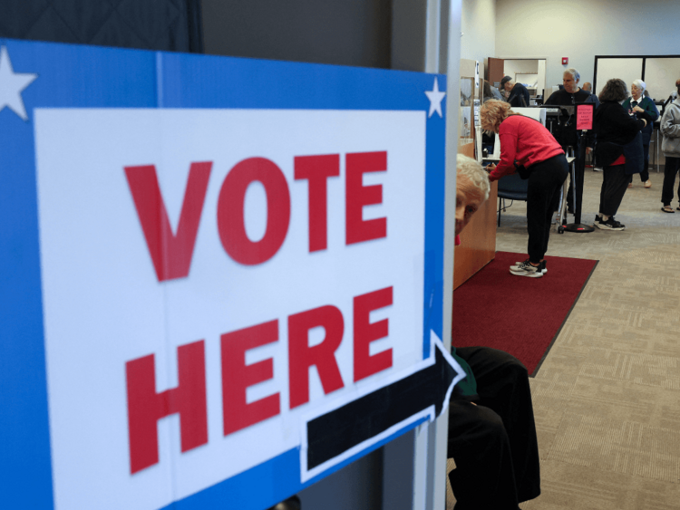 A sign reading "vote here" is displayed in large red, white, and blue letters on a door. People are standing at voting booths inside.