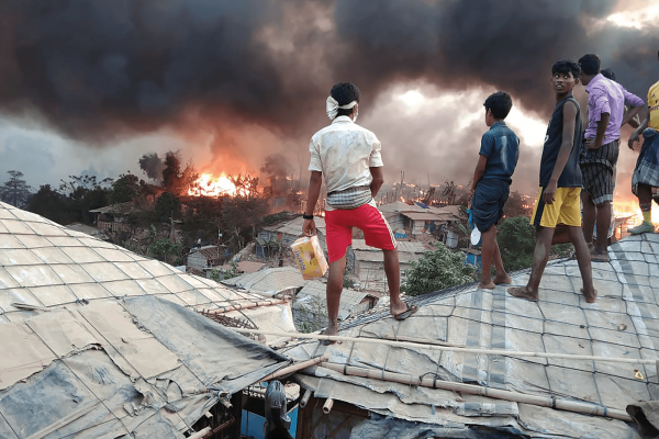 Rohingya youth stand atop tarp roofs as they watch fire spread in the treeline