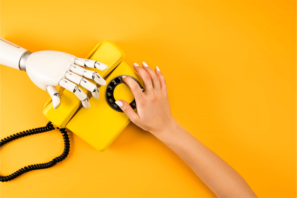 A human hand and a robotic hand both reach for a vintage telephone on a yellow background.