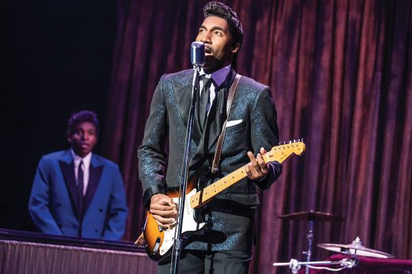 Garrett Turner is dressed in a black suit playing an electric guitar as Ike Turner in the musical 'TINA: The Tina Turner Musical.' A black man in a blue suit is playing the keyboard in the background, where both men are flanked by a purple stage curtain.