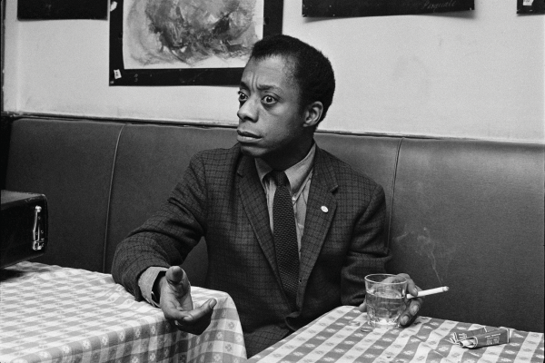 James Baldwin holds a cigarette in one hand and gestures with the other while sitting in a booth