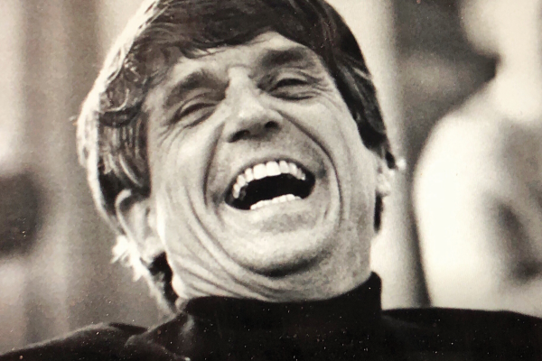 A white man wearing a black turtleneck crinkles his face with exuberant laughter