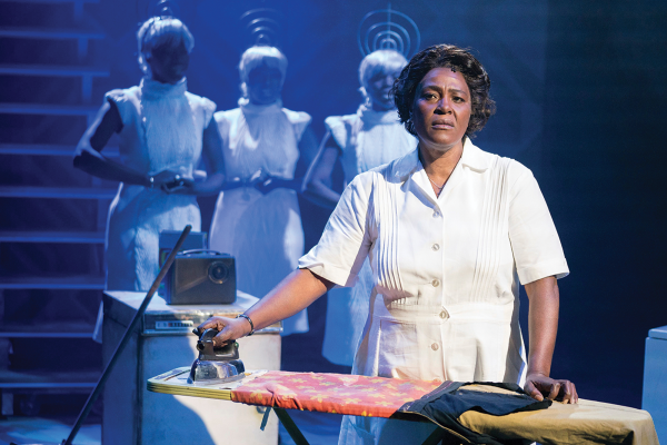 A weary middle-aged Black woman looks up from ironing clothes in a dark room
