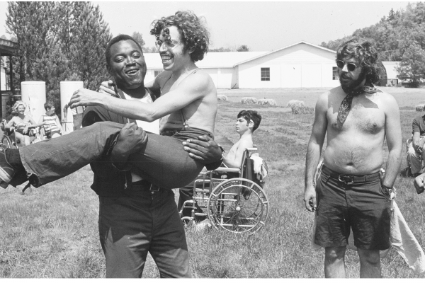 In a scene from the documentary "Crip Camp," a camp counselor is carrying a man who is Disabled. Both are laughing.