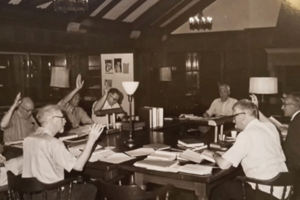 The image is a black and white photo showing a group of old white men sitting around a table with Bibles and other documents, some have their hands raised. 