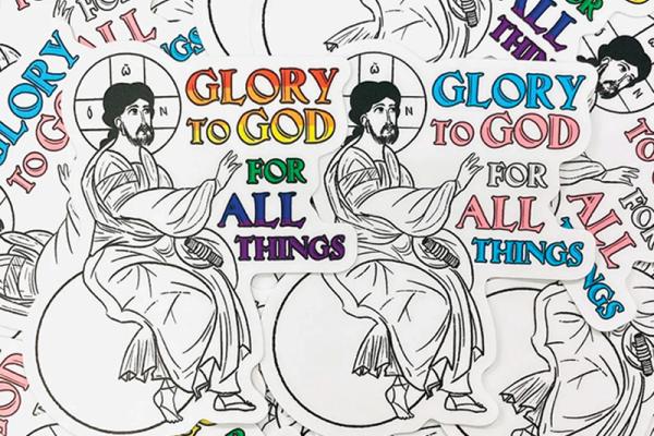 An illustration of Jesus sitting on a globus cruciger as he raises his hand to text that reads, "Glory to God for all things." One version depicts the text in the colors of the LGBT Pride flag and the other the colors of the transgender flag.