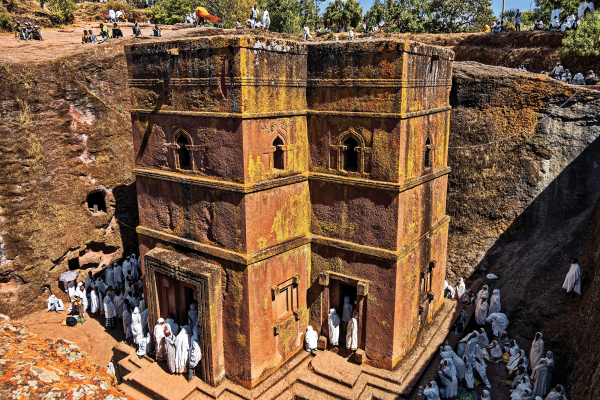 the photo is a image of a red church carved out of rock. The church has pilgrims surrounding it, all of them wearing white