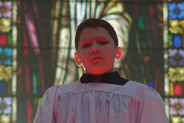 A young boy wearing a white surplice stands before a wall of stained glass