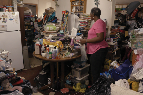 A woman stands in a kitchen with objects piled all over a table and along the walls 
