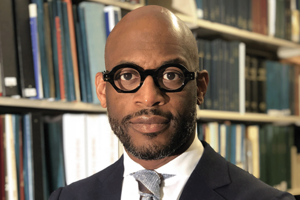 The picture shows a bald Black man with facial hair wearing black glasses in front of a bookshelf, wearing a suit. 