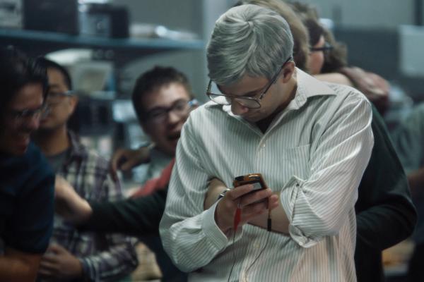A photo from the docufilm ‘Blackberry.’ Actor Jay Baruchel is Mike Lazaridis, co-CEO of Blackberry. He has short gray hair, glasses, and wears a white dress shirt. He glares down at a phone with wires plugged into it. People behind him are cheering.