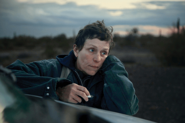 A scene from Nomadland features a woman leaning on the hood of her car.