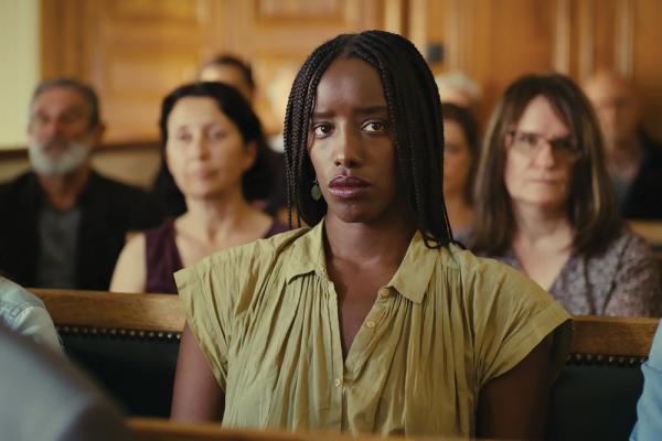 Kayije Kagame plays as Rama in the film ‘Saint Omer.’ She is a Black woman with box braids wearing a creased linen olive-green v-neck dress. She sits in the pews of a court with a crowd of people blurred in the background.