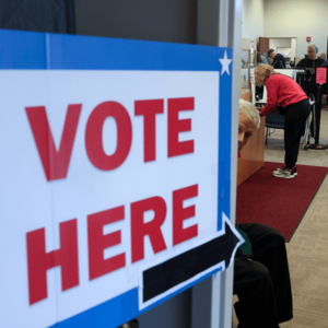 A sign reading "vote here" is displayed in large red, white, and blue letters on a door. People are standing at voting booths inside.