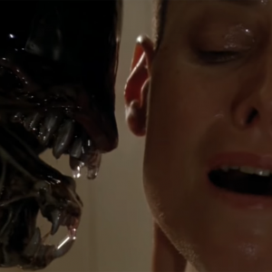 A close-up screenshot from Alien 3; an alien creature with its mouth open is seen from the side, with its teeth next to the shiny face and ear of a white woman who directly faces the camera.