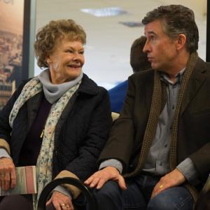 Philomena (played by Dame Judi Dench) and Sixsmith (played by Steve Coogan) sit next to each other in a waiting room. Philomena is wearing a black jacket with a flower-patterned scarf. Sixsmith is wearing a dark brown jacket and blue jeans.