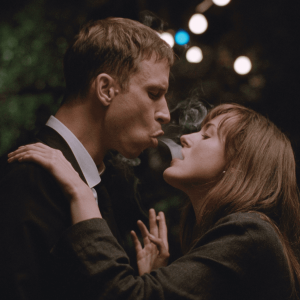 A white man wearing a suit blows smoke into the mouth of a young white woman