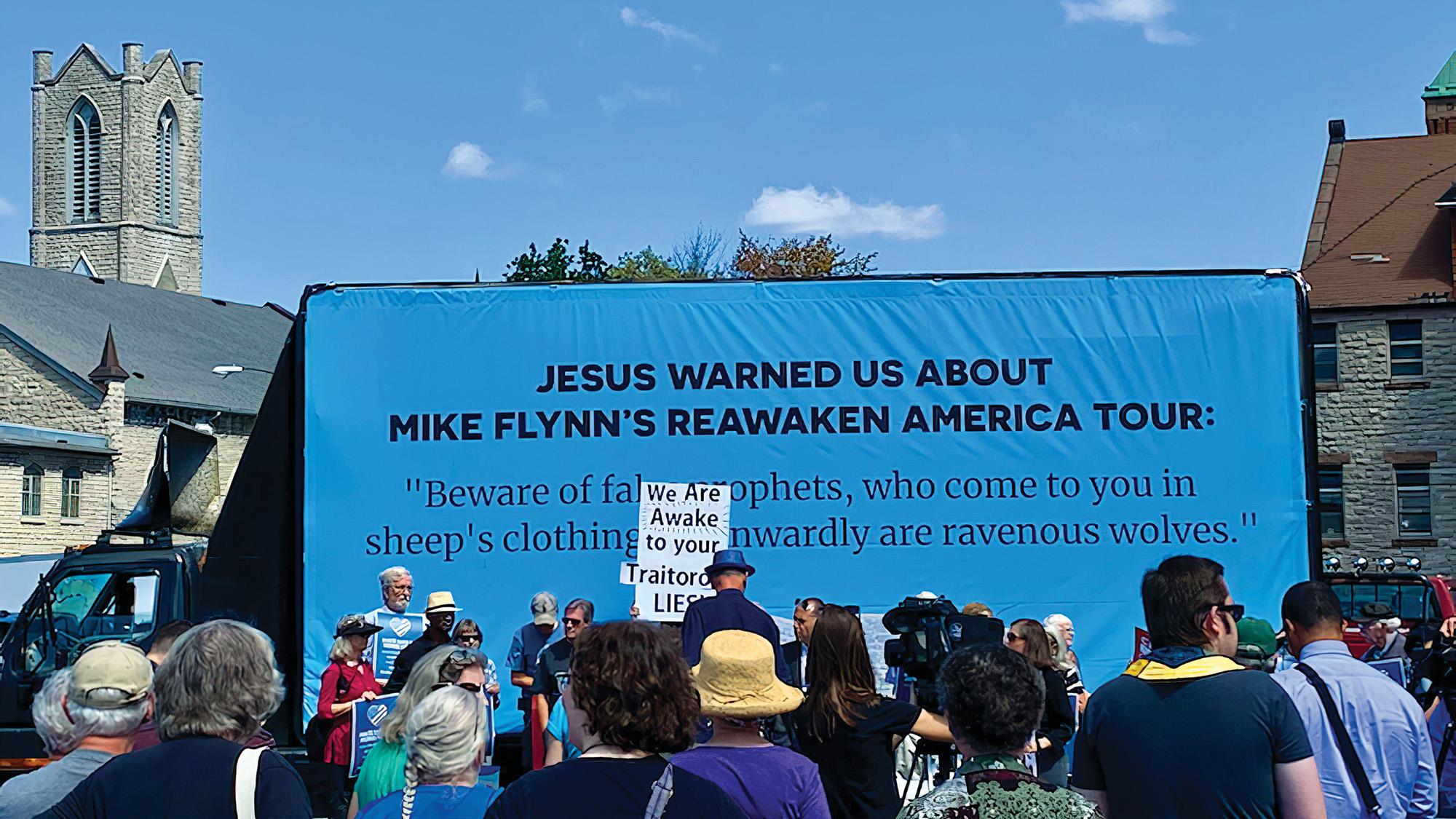 Photo courtesy of Faithful America Photo of a cerulean blue billboard with black text that reads "Jesus Warned Us About Mike Flynn's ReAwaken America Tour." Foreground features a group of people facing the billboard; one protestor's sign reads "We Are Awake to Your Traitorous Lies"