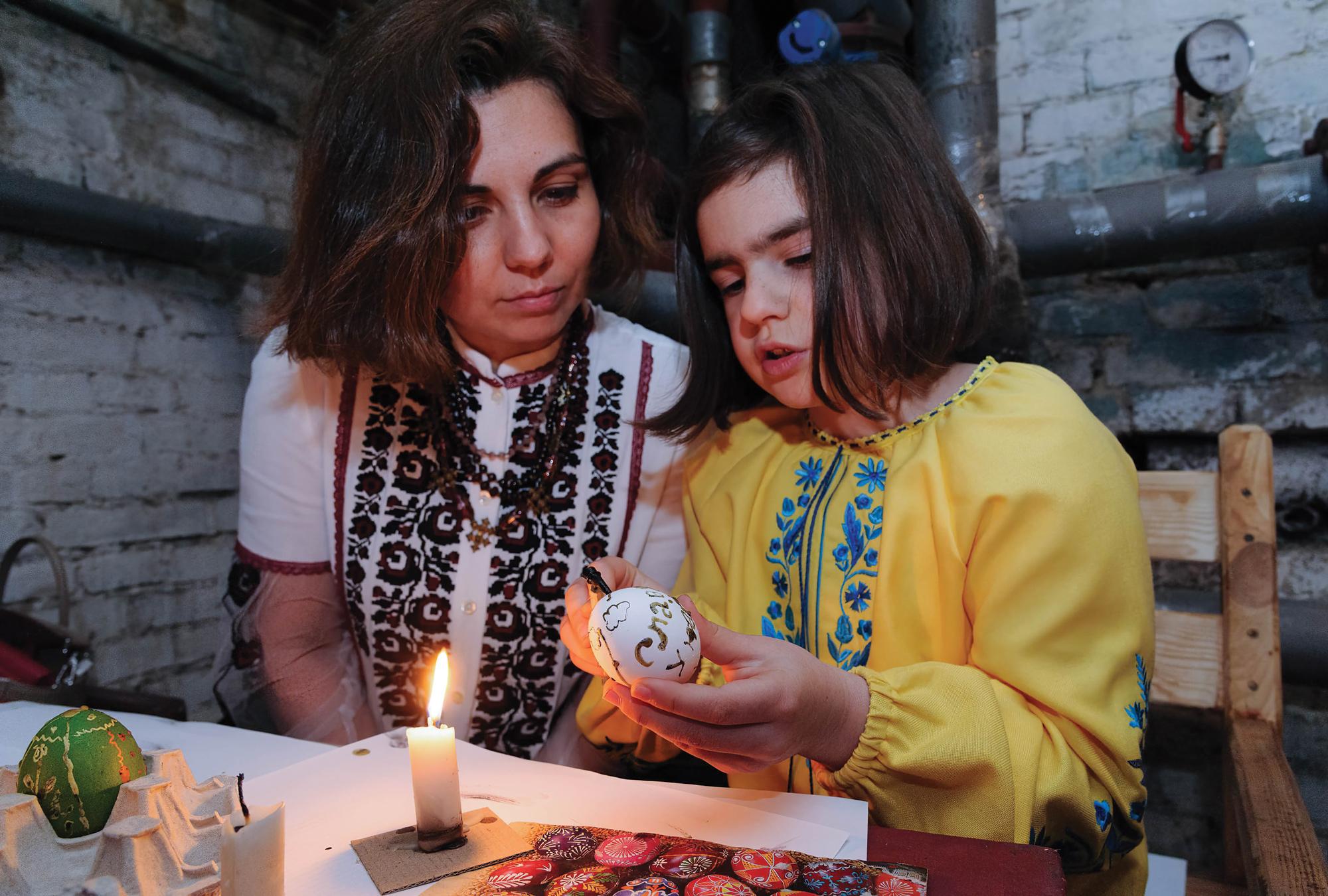 A Ukrainian woman and girl are sitting together as they paint an Easter egg.