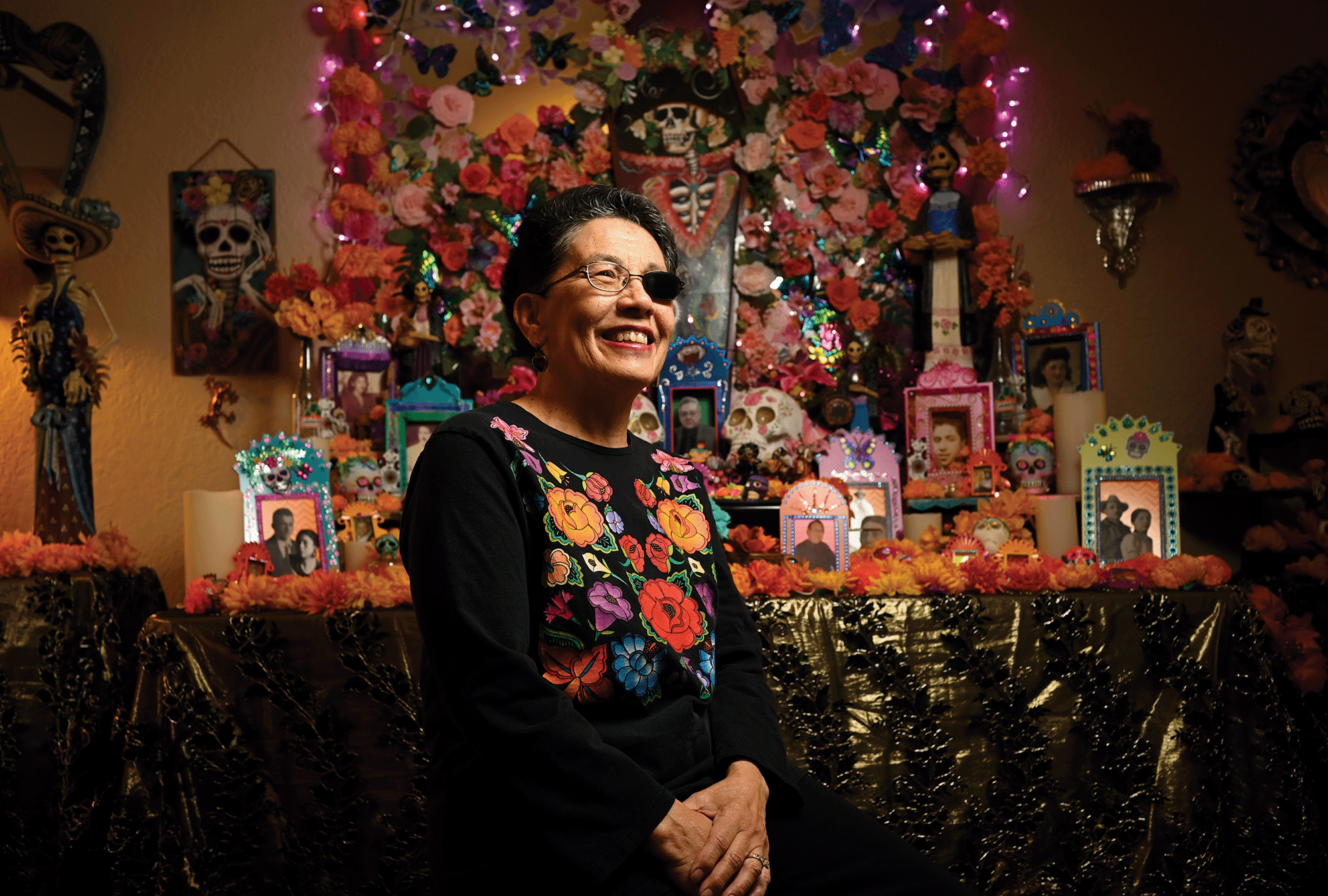 Hyoung Change/The Denver Post A smiling woman with short hair and an eye patch stands in front of a colorful Dia de los Muertos altar which has pictures of deceased loved one and flowers.