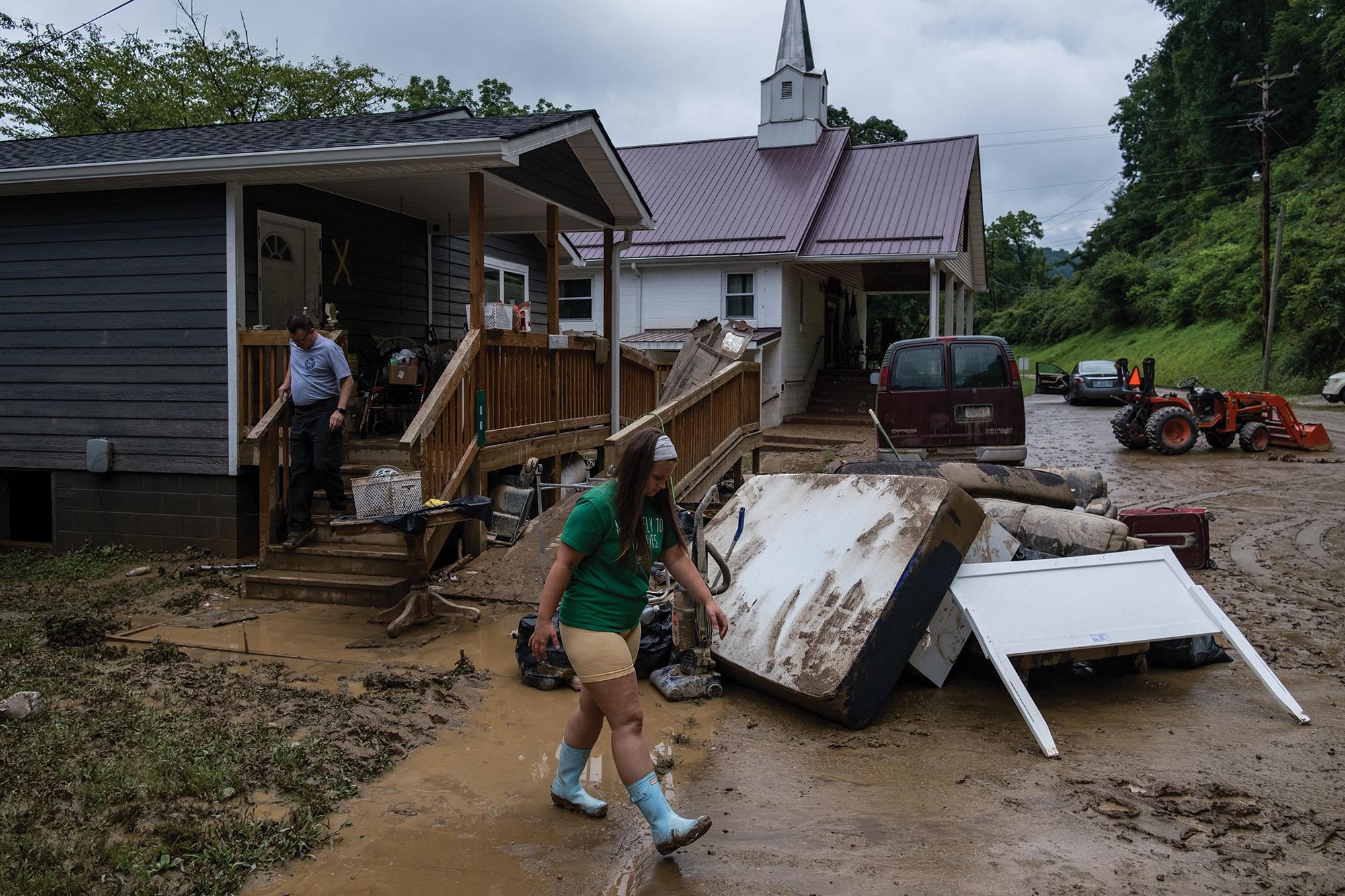 Seth Herald / AFP via Getty Images Two people walk out of a house surrounded by muddy streets and ruined furniture from a flood.