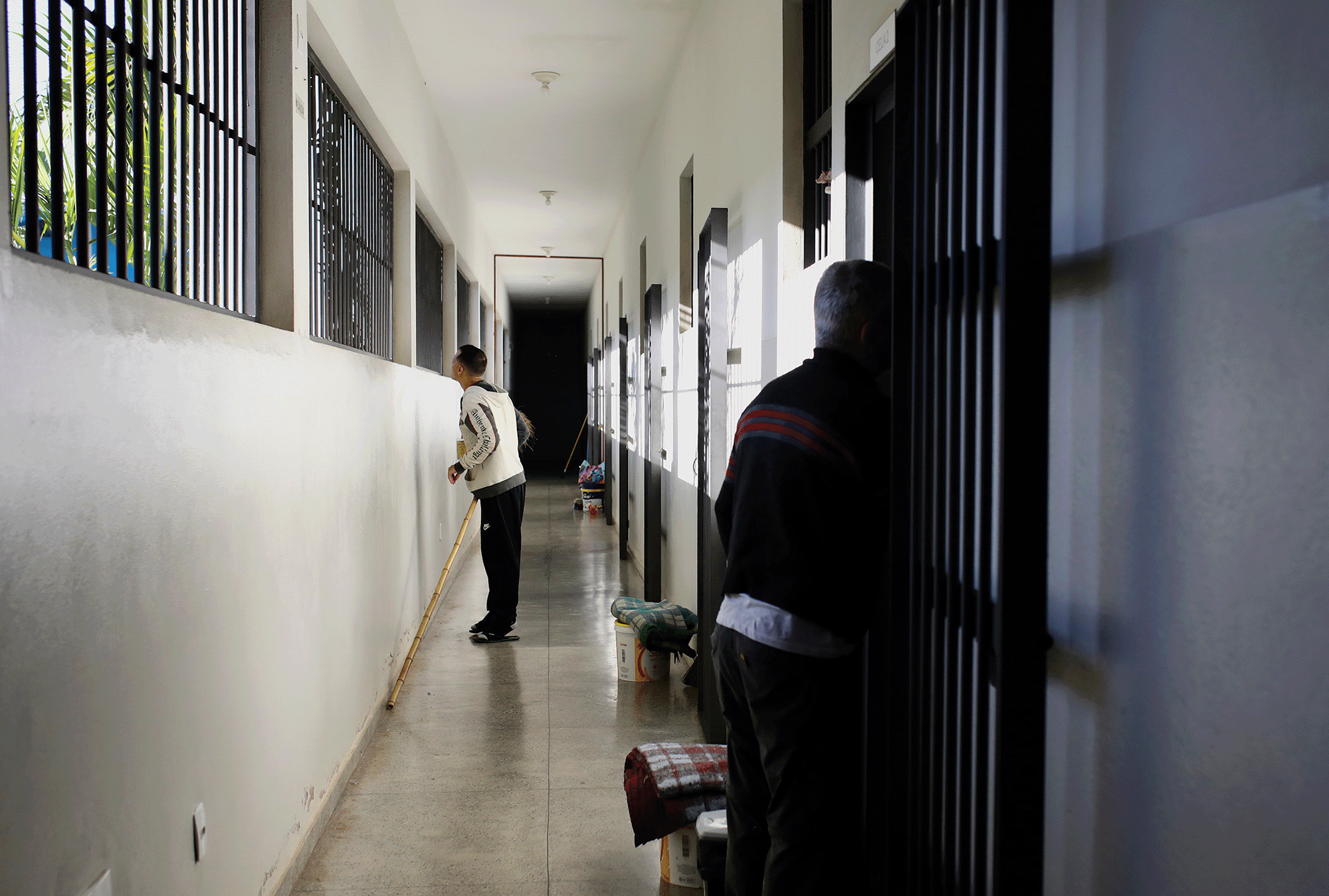 Apolline Guillerot-Malick Pictured is a hallway with white walls and barred windows. There is a man in a black pants and a white sweatshirt looking out of the window, with another man standing down the hall looking at him.