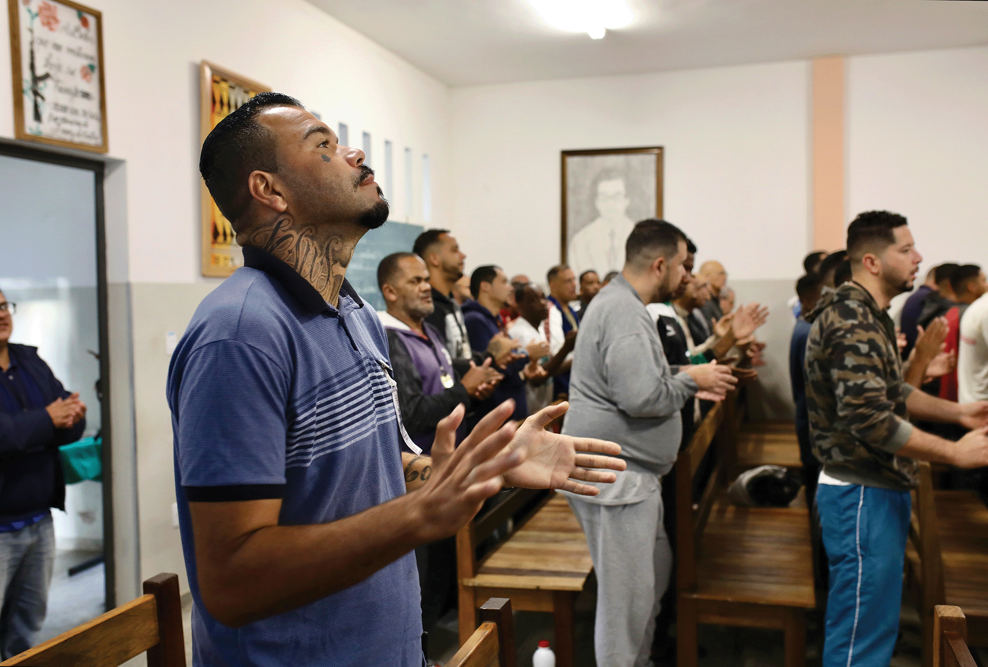 A room is filled with men standing up, clapping their hands together, singing a worship song. The man closest to the camera is wearing a blue shirt and is looking towards the ceiling. 