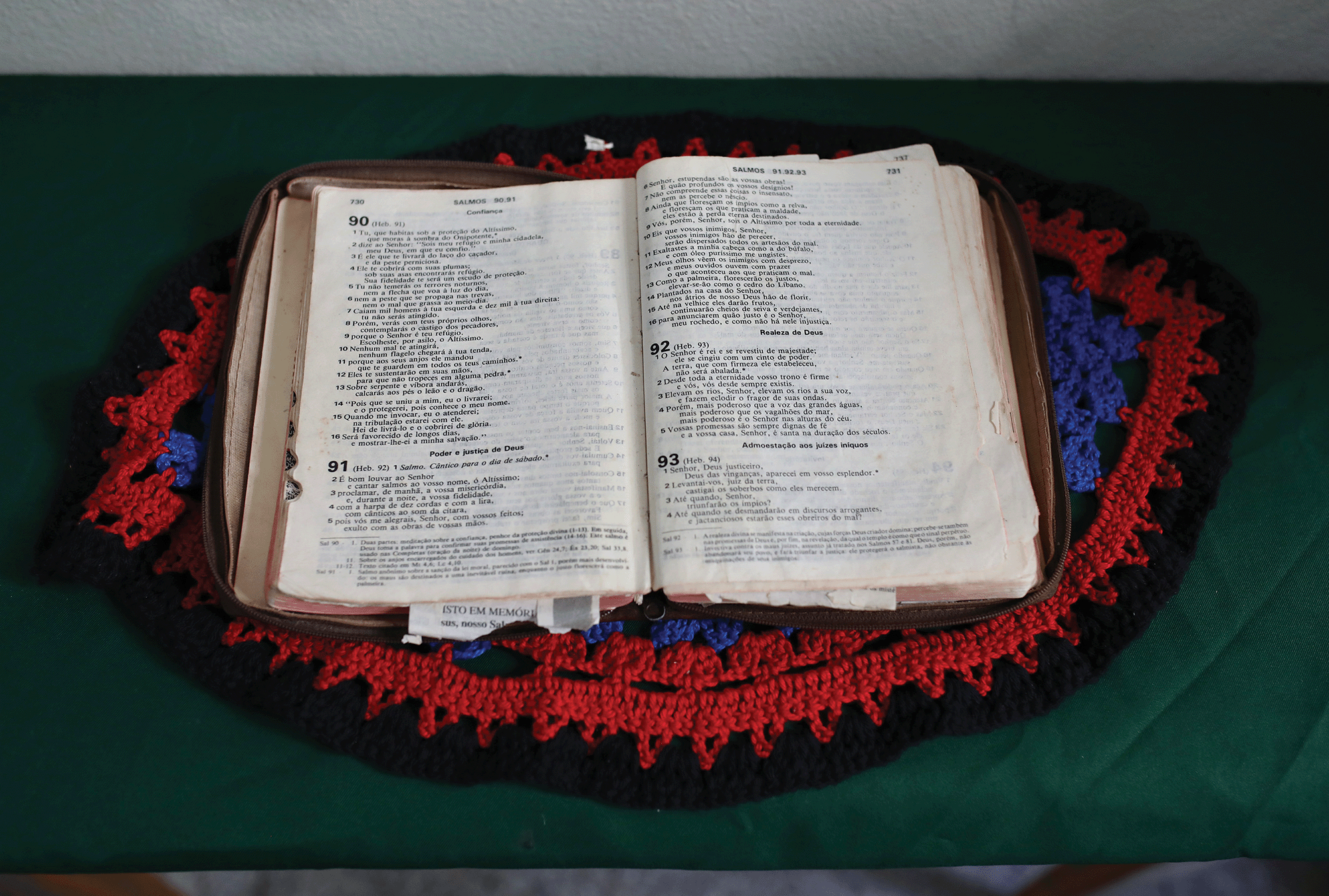 An open Bible in Portuguese sits on a red and blue crocheted placemat on a green tablecloth. 