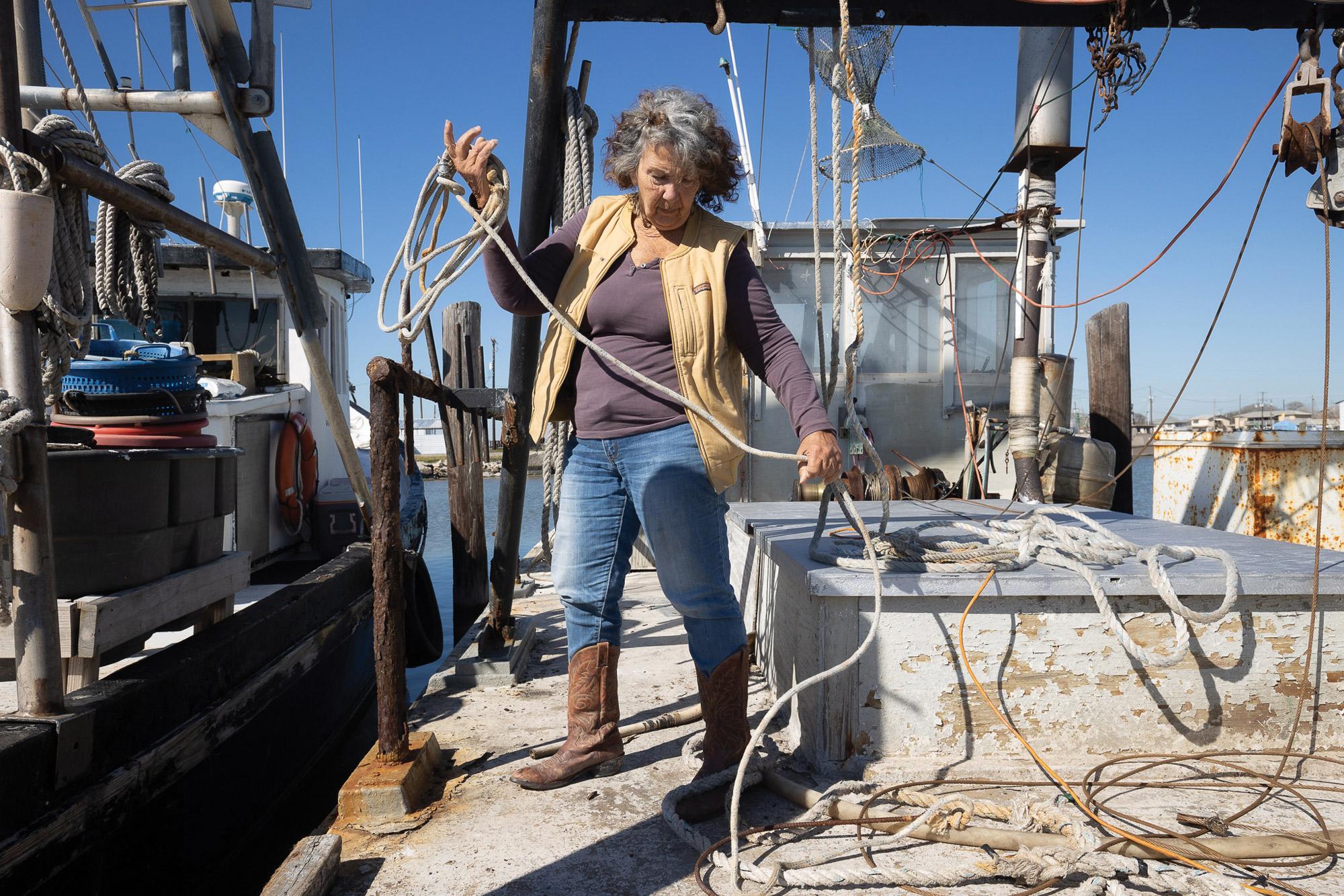 Goldman Environmental Prize The author of this piece, Diane Wilson, is wearing a tan vest with a purple long-sleeve shirt, jeans, and brown boots. She's rolling up rope on a boat, which is docked next to another one to the left.