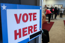 A sign reading "vote here" is displayed in large red, white, and blue letters on a door. People are standing at voting booths inside.
