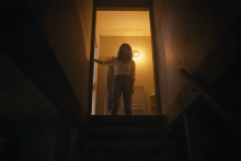 A back-lit woman stands at the top of basement stairs, looking down.