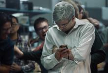 A photo from the docufilm ‘Blackberry.’ Actor Jay Baruchel is Mike Lazaridis, co-CEO of Blackberry. He has short gray hair, glasses, and wears a white dress shirt. He glares down at a phone with wires plugged into it. People behind him are cheering.