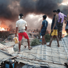 Rohingya youth stand atop tarp roofs as they watch fire spread in the treeline