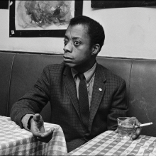 James Baldwin holds a cigarette in one hand and gestures with the other while sitting in a booth