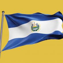 A picture of El Salvador's blue and white national flag, flying from a flag pole against a yellow backdrop.
