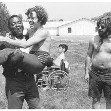 In a scene from the documentary "Crip Camp," a camp counselor is carrying a man who is Disabled. Both are laughing.