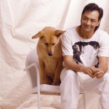A photo of Rich Mullins in a white t-shirt and white pants. He's sitting on a white chair with a white sheet draped in the backdrop. He's leaning forward on his knees with a grin on his face. A dog with tan-colored fur sits to his right on the same chair.