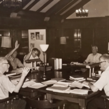 The image is a black and white photo showing a group of old white men sitting around a table with Bibles and other documents, some have their hands raised. 
