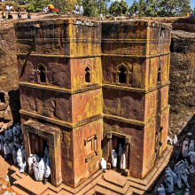 the photo is a image of a red church carved out of rock. The church has pilgrims surrounding it, all of them wearing white