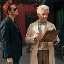 The photo shows two men, one who is an angel and dressed in lighter colors, and another who is a demon dressed in black. The angel is looking at a clipboard and the demon is just standing there. 