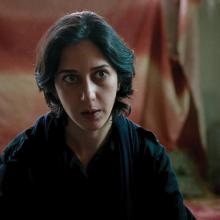 A photo of actress Zar Amir Ebrahimi as fictional journalist Arezoo Ramimi in the film 'Holy Spider.' She is cast against a red flag in the background and staring just off camera at something.