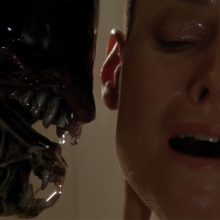 A close-up screenshot from Alien 3; an alien creature with its mouth open is seen from the side, with its teeth next to the shiny face and ear of a white woman who directly faces the camera.