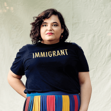Aline Mello stares seriously at the camera wearing a multicolored skirt and a t-shirt that says "Immigrant"