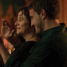A son embraces his mother from behind, who lift up their hands together to clap.