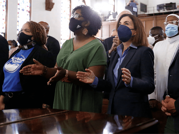 A diverse crowd of people stand in a church wearing masks during a vigil.