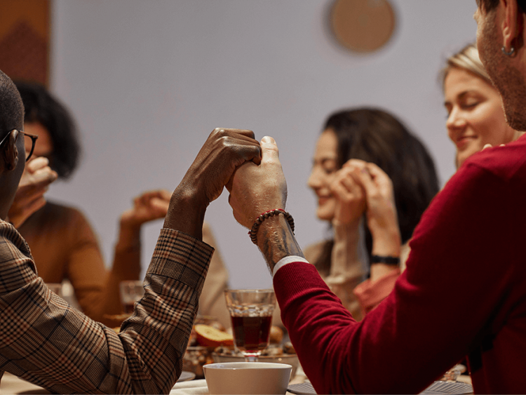 A multiracial group of people hold hands in prayer at Thanksgiving dinner.