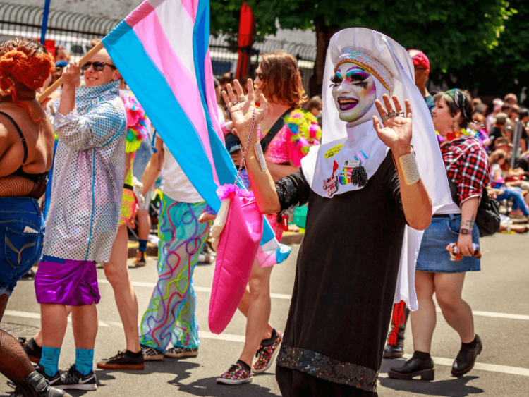 A person wearing heavy drag makeup and a black Catholic habit waves in a pride parade.