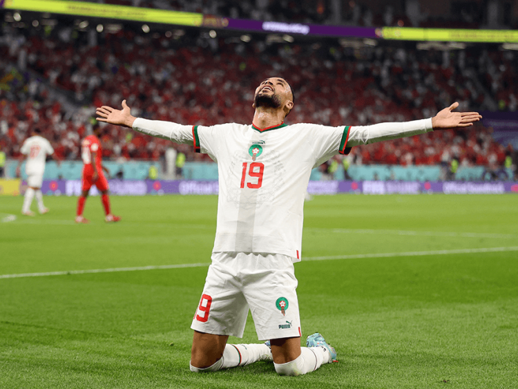 A soccer player kneels on the field with arms outstretched. Behind him, fans are crowded in stadium seating.