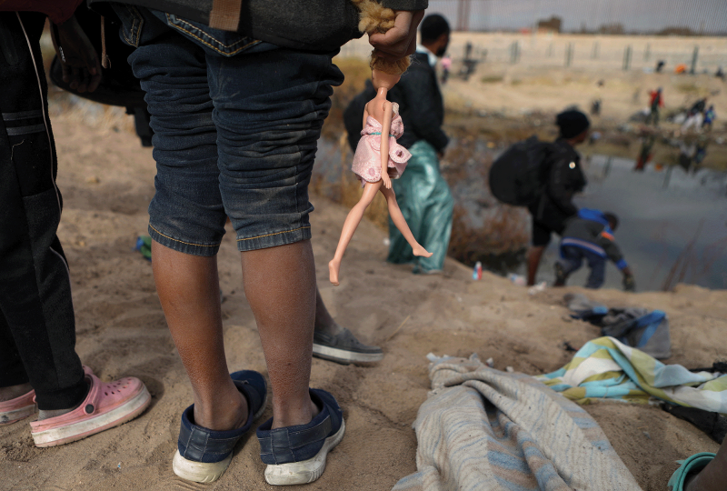 The picture shows the legs of a girl standing on the bank of the Rio Grande, holding a Barbie doll by its hair. 