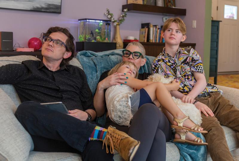 A family photo of trans Christian Sid High's family sitting on a couch in their living room as they look off to the side. Sid's dad sits on the left; Sid's mom sits in the center with his younger sister in her lap; Sid sits on the right.